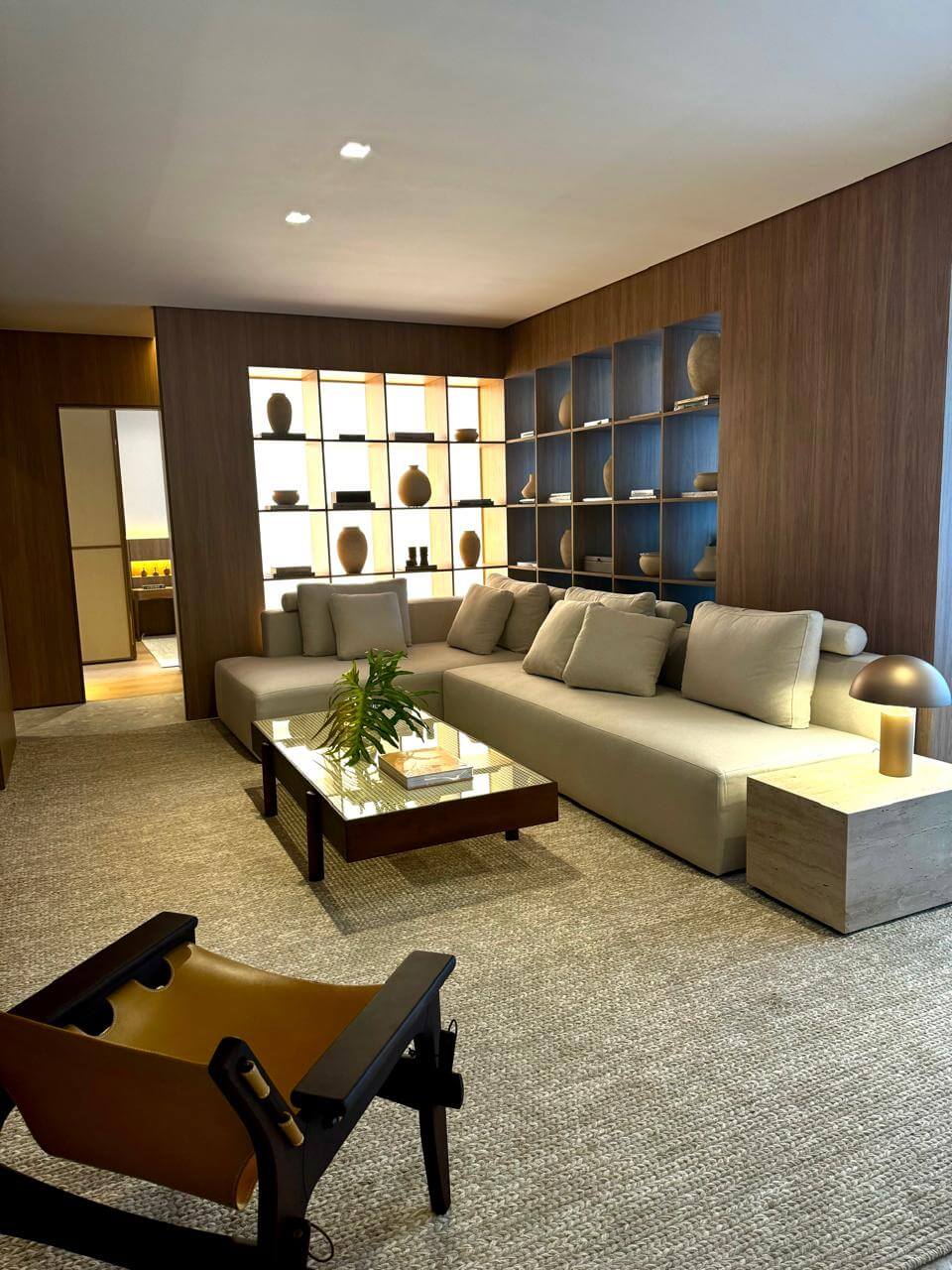 Novo luxo no mercado imobiliário: experiência e bem-estar 2 Living com nichos iluminados por luz embutida e abajur sobre mesa lateral de travertino, em tons quentes