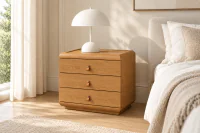 Mesa de Cabeceira Elora em acabamento Carvalho Nobre, com três gavetas e puxadores em couro Caramelo, ambientada em quarto contemporâneo elegante, com luminária branca sobre o tampo, cortinas claras e decoração em tons neutros.