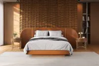 Duas Mesas de Cabeceira Redondas Avila em Lâmina Natural de Cinamomo com tingimento Castanho e estrutura metálica Champagne Metalizado, posicionadas ao lado de cama estofada caramelo em quarto de casal contemporâneo, com painel de madeira, tapete claro e decoração sofisticada.